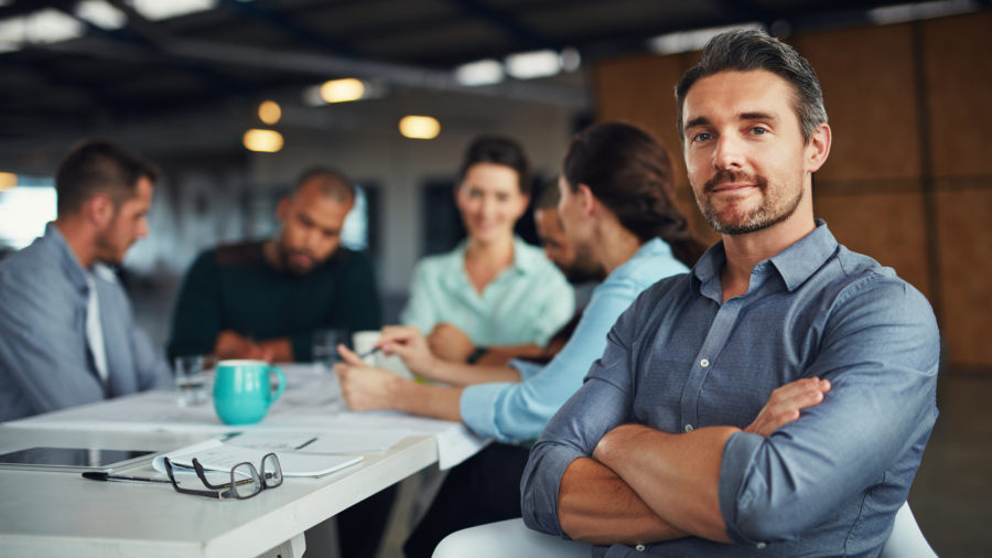 Confident business professional seated at a meeting table with team collaborating in the background