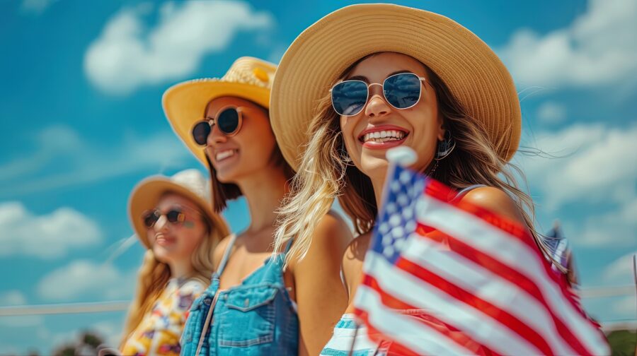 Three smiling women in sun hats and sunglasses celebrate July 4th outdoors, holding an American flag under a bright blue sky.