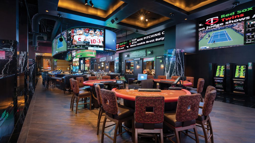 Sportsbook interior with gaming tables, bar seating, and large overhead screens displaying baseball and tennis broadcasts.