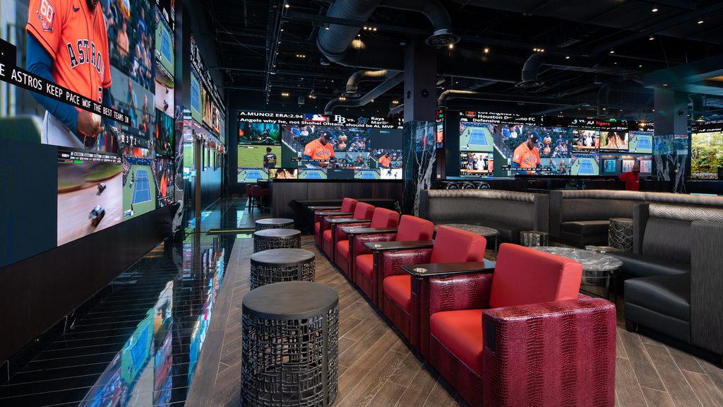 Sports lounge interior featuring red armchairs, booth seating, and large video walls displaying multiple live sporting events.